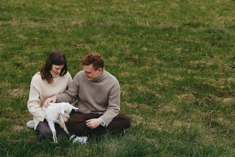 Lifestyle couple gently pets a white goat kid in an outdoor grass field, cozy sweaters, natural candid moment, warm bonding and calm affection, a simple rural scene of care and joy.