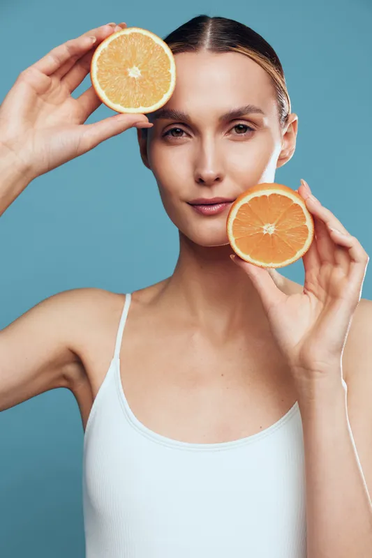 A young woman in a white tank top holds two halves of a grapefruit near her face against a blue background. The image represents healthy lifestyle, skincare, and natural beauty concepts.