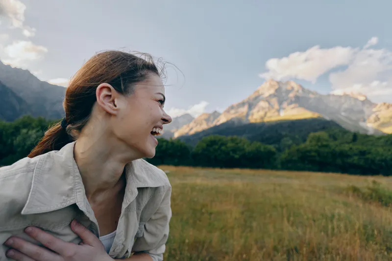 A smiling woman in casual clothes enjoys the outdoors with a mountain and green forest in the background under a partly cloudy sky. Lifestyle and nature concept.