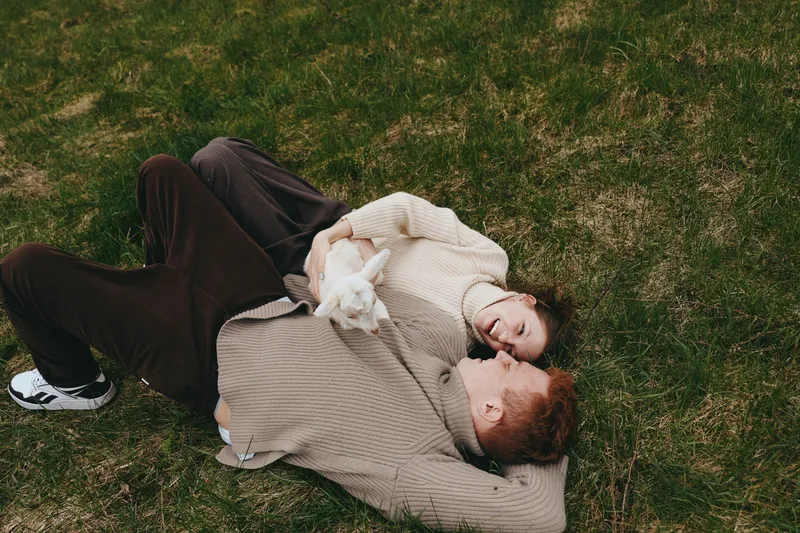 Couple lies on a grassy field, wrapped in cozy sweaters, sharing a tender moment outdoors with a small dog, conveying warmth, relaxation, and affectionate companionship. Their casual posture and soft