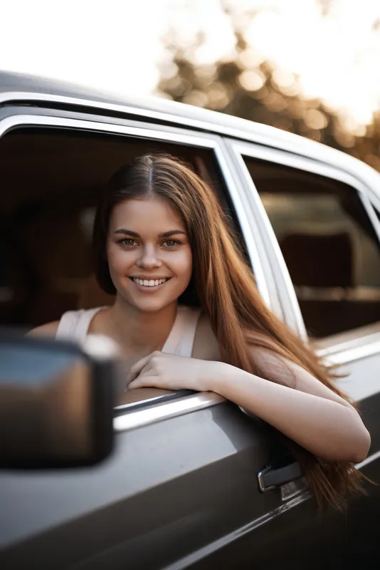 Young girl smiling from car window in sunny setting, showcasing joy, happiness, and a carefree attitude with warm color tones and natural background.