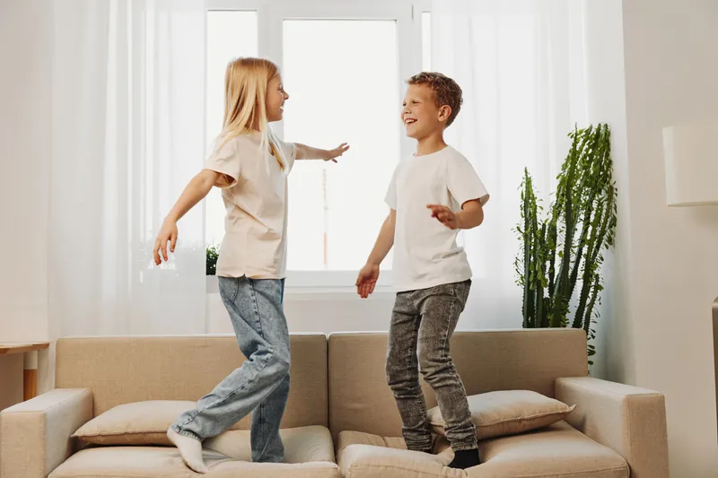 Two happy children jump on a beige sofa in bright living room, enjoying playful moment and carefree fun together at home.