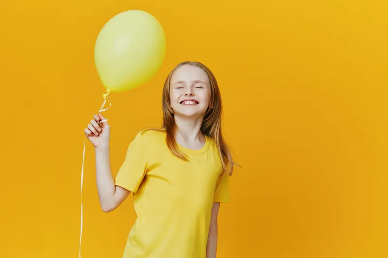 A cheerful girl with long hair in a yellow t shirt happily holding a yellow balloon against a bright yellow background, embodying joy and happiness