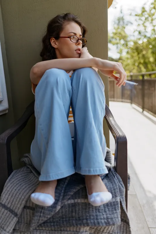 A contemplative woman sits on a balcony in denim jeans and white socks, leaning forward with arms wrapped around knees, enjoying a quiet outdoor moment.