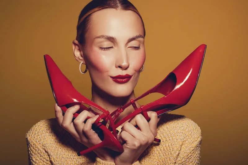 Young woman with closed eyes holds red high heel shoes near face, wearing yellow sweater and gold earrings. Concept of fashion, style, elegance, and beauty with vibrant colors.