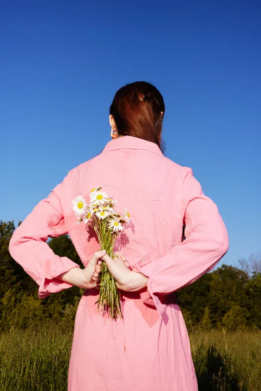 Woman holding flowers behind her back, wearing a pink dress, smiling in a sunny field, representing joy, nature, and beauty in springtime