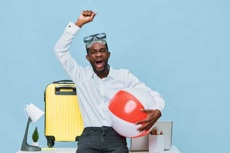 Happy cheerful man with dark skin celebrating vacation travel success holding beach ball and wearing sunglasses, standing at modern office desk with yellow suitcase, travel accessories, on bright blue