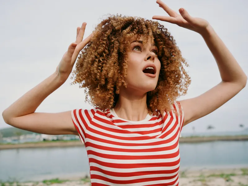 A joyful woman with curly hair raises her arms in a candid outdoors portrait, wearing a red and white striped shirt, beside a calm water body and distant shoreline.