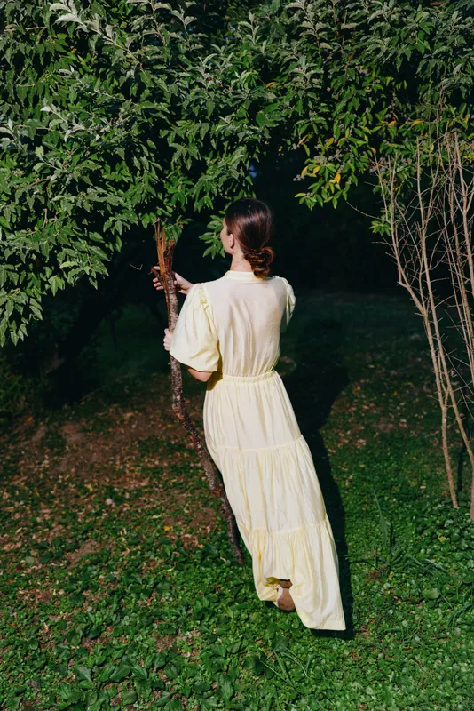 Woman in a pale yellow dress walks away through a tranquil garden at night among trees, capturing a calm cinematic moment in soft moonlight.