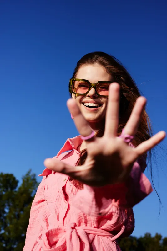Happy young woman in pink outfit enjoying sunny outdoor day, smiling playfully against a clear blue sky with vibrant green trees in the background