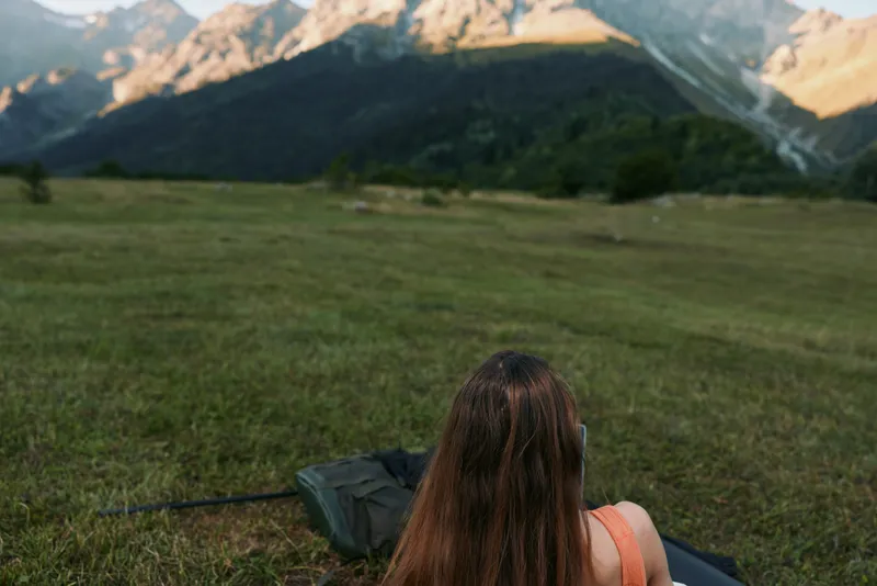 A person with long hair sits on a grassy hillside, facing distant mountains, with a backpack nearby. The scene conveys solitude, outdoor exploration, and a moment of reflection in nature.