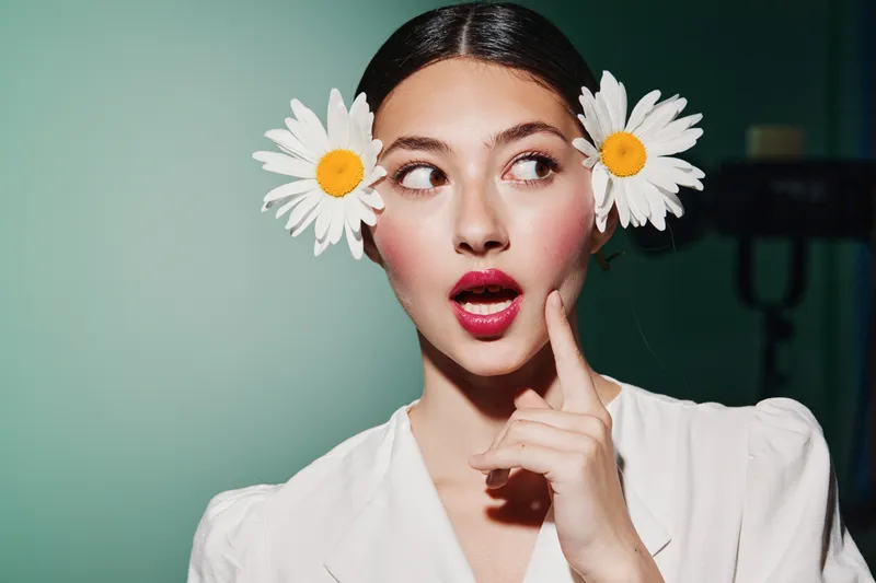 Young woman with flowers in her hair shows surprise with pink lips and finger near her cheek in bright studio light. Concept of beauty creative fashion and floral style.