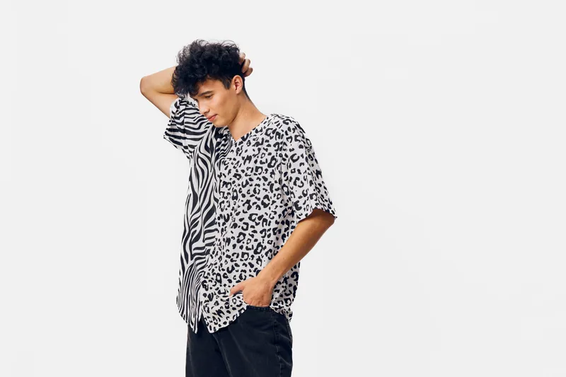 Young man in patterned black and white shirt standing with hand on head against white background, casual style and thoughtful expression, monochrome outfit.