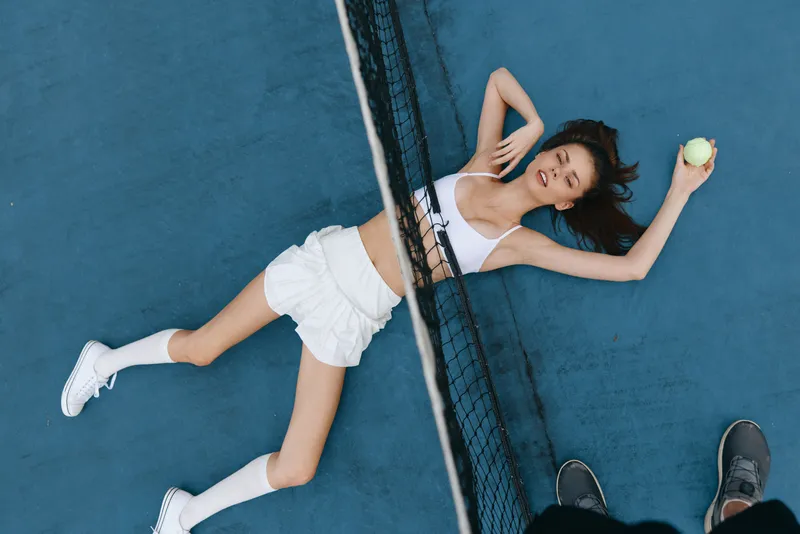A young athletic woman with a joyful expression sprawled on a blue tennis court, wearing a white crop top and shorts, holding a tennis ball, showcasing the sport s energy