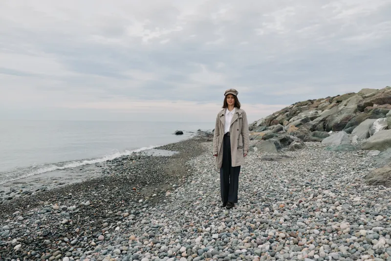 Woman in beige coat and hat stands on rocky beach near calm sea under cloudy sky. Concept for autumn, travel, nature, and outdoor lifestyle with peaceful mood and neutral colors.