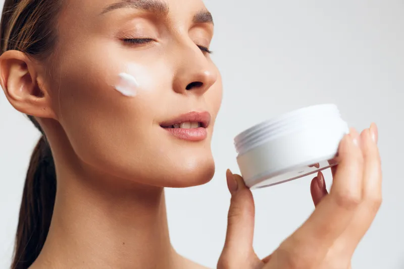 Young woman with smooth skin gently applies moisturizer on her cheek in a close-up shot, white studio background, skincare routine, self care, relaxing and focused mood