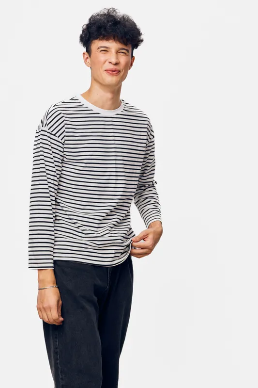 young man in casual striped long sleeve shirt smiling playfully with closed eyes, standing in front of white background, wearing black pants