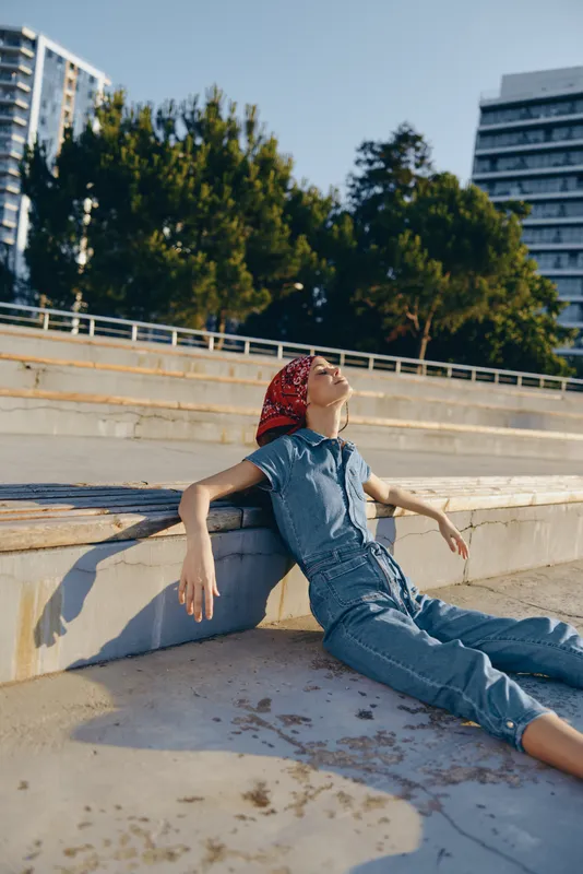 relaxed woman wearing denim jumpsuit and red bandana sitting outdoors on concrete steps under warm sunlight with urban background and green trees.
