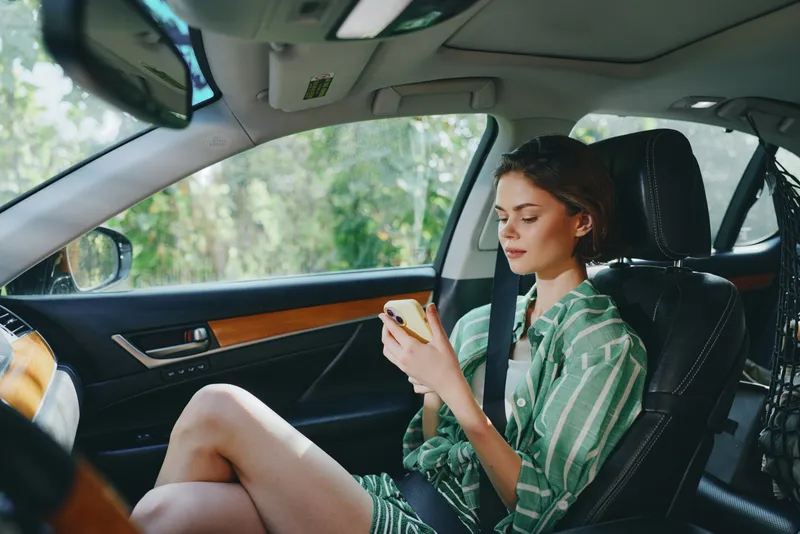 Young woman sits in the car, wearing a casual green striped shirt, focused on her smartphone.