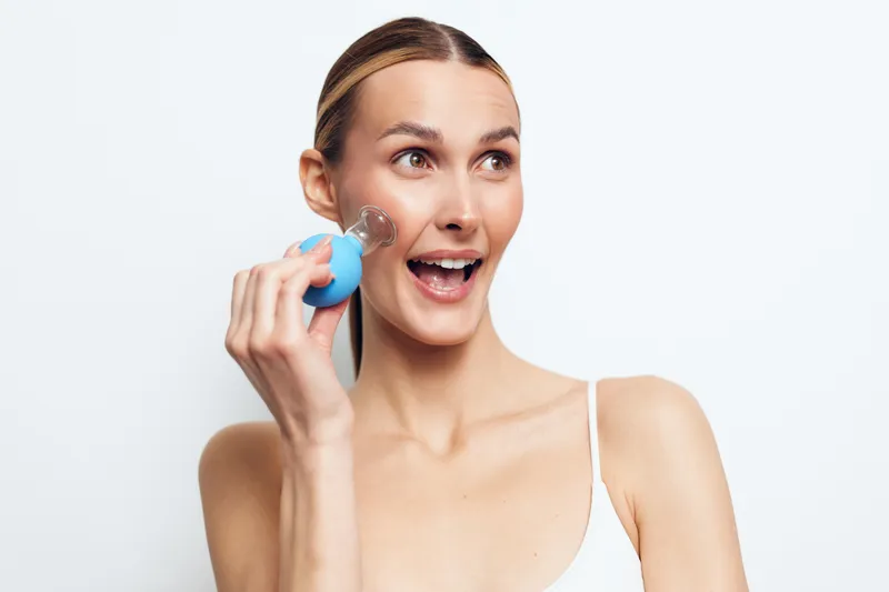 Young woman with blonde hair uses a blue facial cleansing brush on her cheek while smiling and looking slightly to the side against a plain white background.
