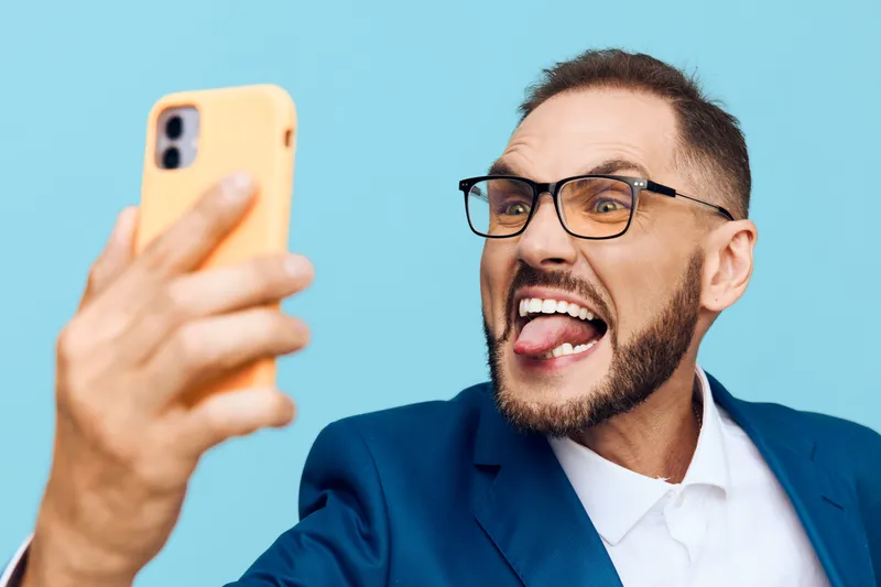 A professional man in a tailored suit takes a lively selfie, displaying confident, excited emotions while the isolated blue background highlights his dynamic presence.