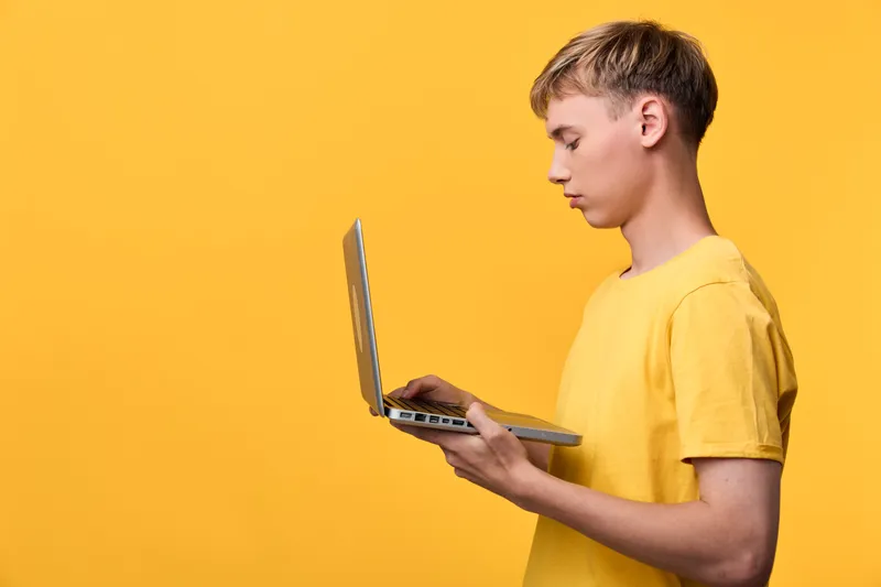 Boy holding a laptop in a bright yellow studio, focused on screen for learning, technology, and education, modern device for online study and exploration