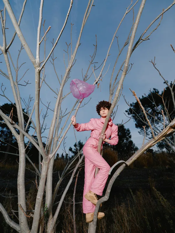 Woman in pink jumpsuit climbs a bare tree in an open field, holding a pink balloon, showcasing playful adventure and bold fashion against a clear blue sky and vast horizon.