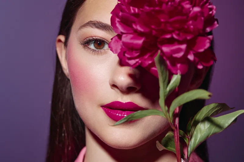 Young woman with bright vibrant makeup and pink lips holds a large dark pink flower covering half her face, with purple background. Creative beauty and fashion concept.