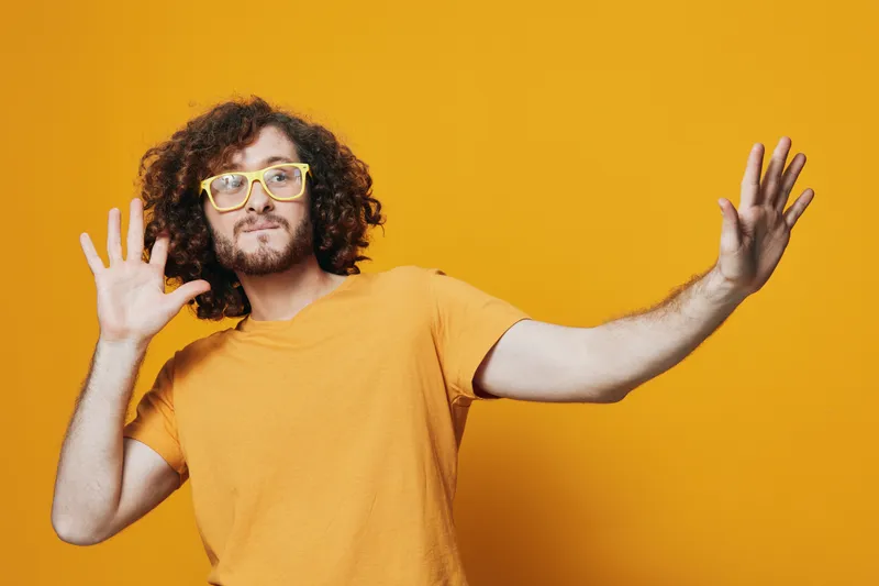 Happy young man with curly hair wearing a yellow t shirt and glasses, expressing joy with hands raised against a vibrant yellow background