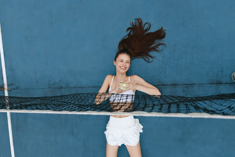 Young woman smiling while holding a tennis ball, wearing athletic clothing, against a blue wall, showcasing a vibrant and energetic sport concept