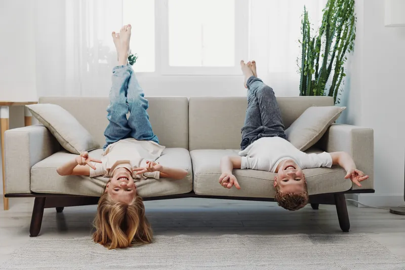 Two happy children in casual clothes lie upside down on a sofa in a bright living room, enjoying playful and carefree moments together.