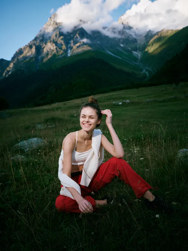 Woman sits on a grassy meadow in a mountain landscape, smiling softly, wearing a white top and red pants, enjoying nature and fresh air in a scenic alpine setting
