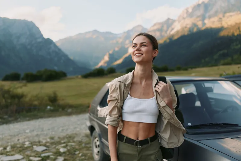 A smiling woman stands beside a SUV in a valley with towering mountains, wearing a white crop top and light jacket. A relaxed travel moment showcasing outdoor lifestyle.