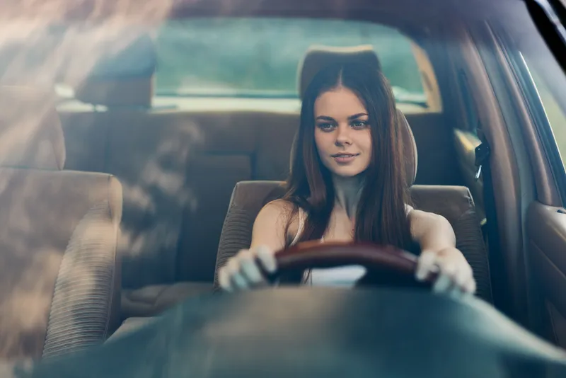 Young woman driving a car, focused expression, with long dark hair, casual attire in a modern interior, evoking freedom and independence.