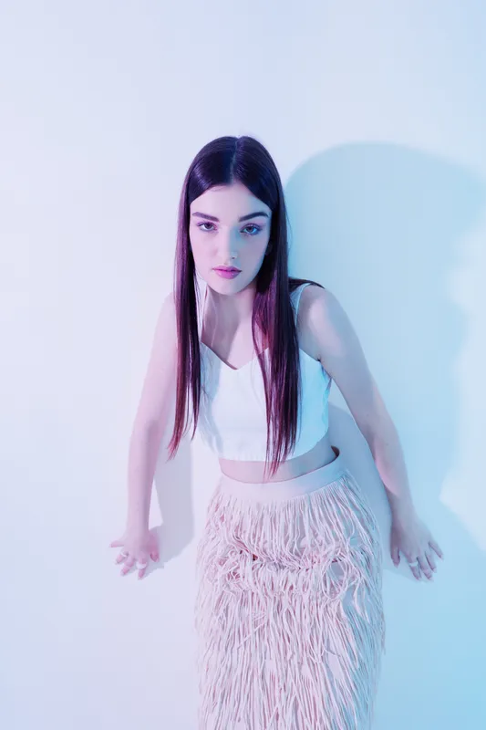 Young woman with long dark hair wearing a white crop top and fringed skirt poses against a pastel background. Concept of beauty fashion style and modern trends.