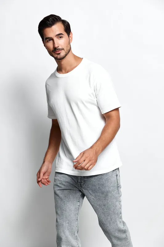 Stylish young man in casual white t-shirt and gray jeans walking with a smile, looking confident and relaxed against a light background. Fashion lifestyle concept.