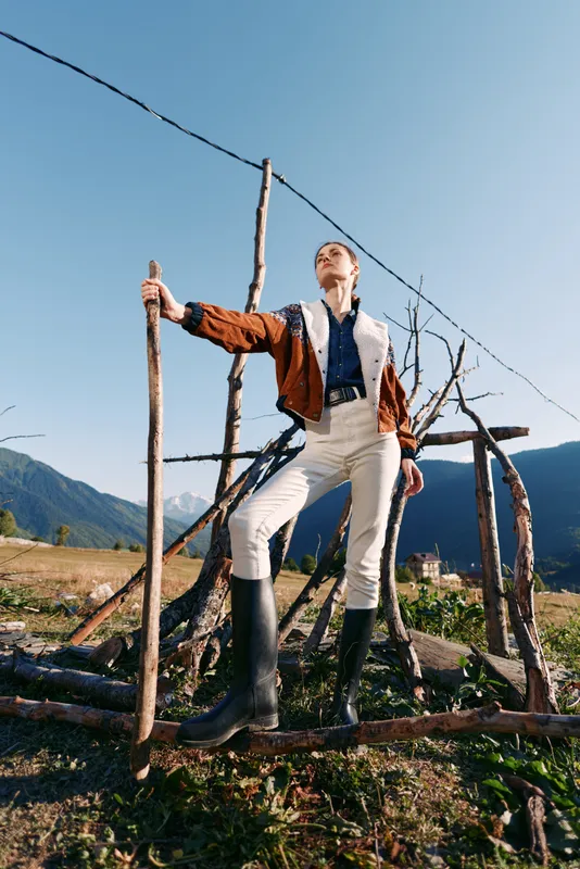 Woman hiking in mountains with a walking stick and rubber boots, standing by a rustic fence in the countryside wearing a brown jacket and white trousers, outdoor portrait and nature scene.