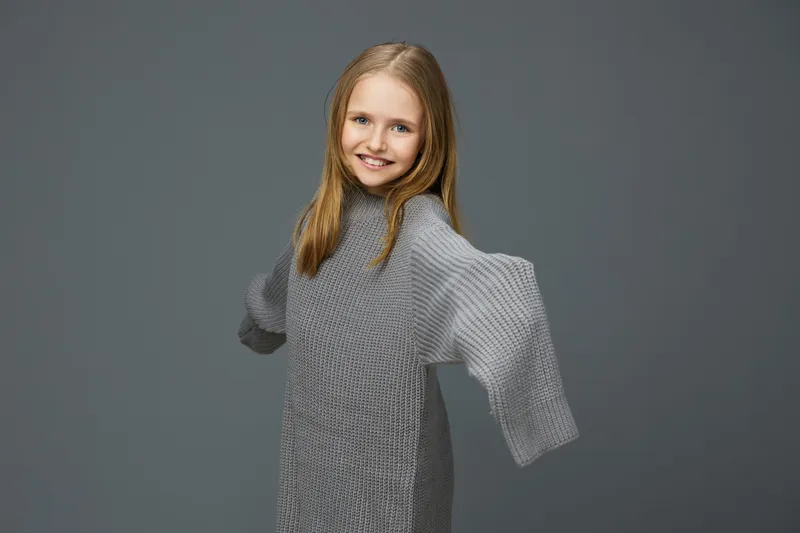Smiling girl in a cozy oversized sweater against a muted grey background, exuding warmth and joy in a contemporary fashion style