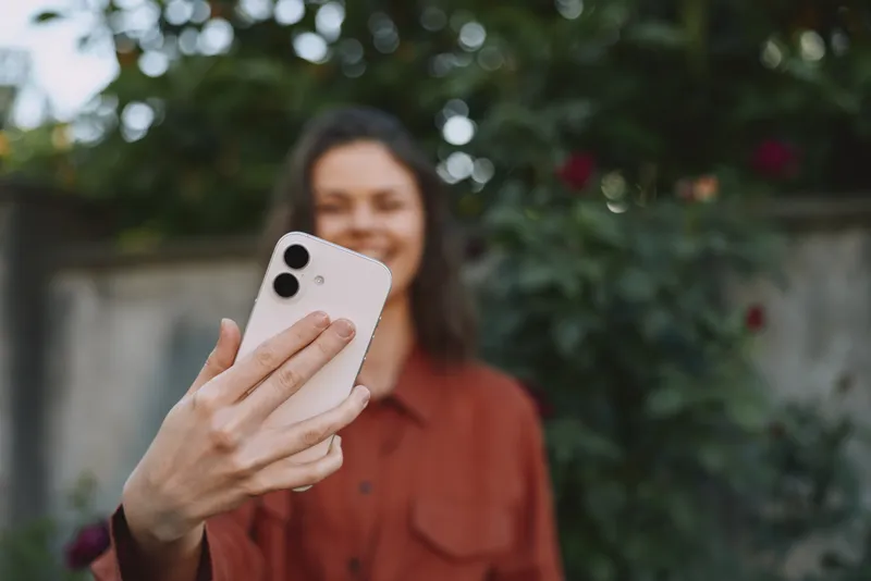 A woman in a casual shirt takes a selfie in a sunny outdoor garden, smiling into the phone. A lifestyle concept focusing on everyday moments and connectivity