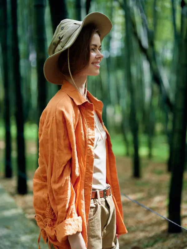 Woman portrait with hat smiling in a bamboo forest, nature outdoors travel scene. Casual orange shirt, relaxed profile and serene green background with soft sunlight.