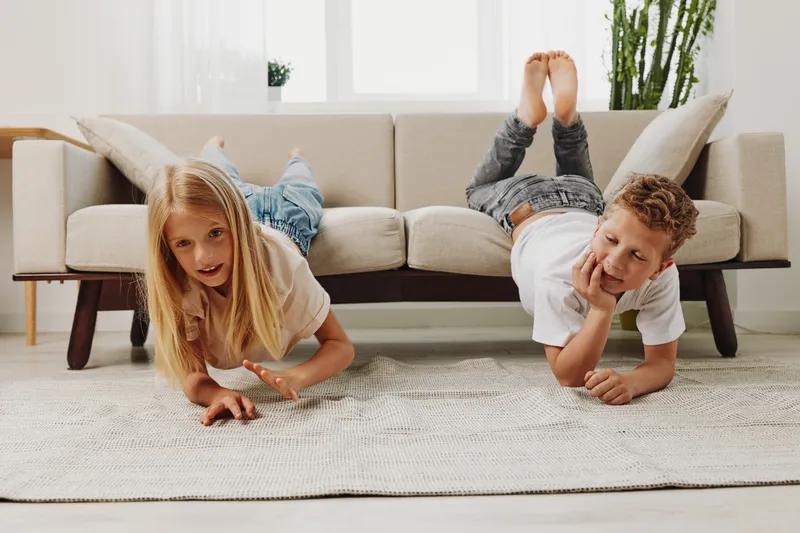 Playful children lying on floor in bright living room near sofa, enjoying leisure time together. Cozy home interior with natural light, carpet, and relaxed childhood moments.