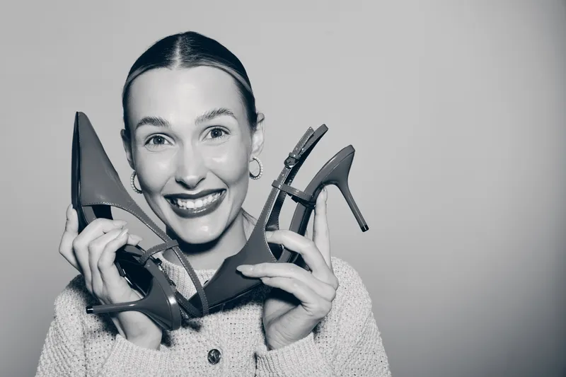 Smiling young white woman with fair skin holds two high heel shoes near face, wearing sweater and earrings. Concept of fashion, style, and joyful female model portrait.