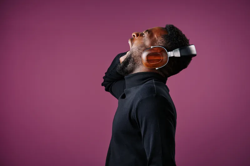 Young Black man enjoying music with headphones on a purple background, expressing joy and freedom in a stylish black outfit while showcasing a modern lifestyle