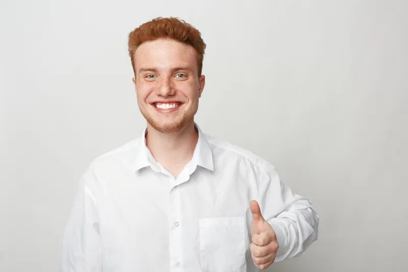 Smiling young man with red hair wearing white shirt shows thumbs up gesture. Portrait on light background. Concept of positivity, approval, confidence, and friendly communication.