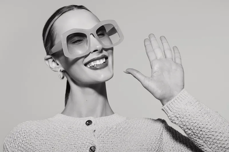 Smiling white woman with light skin and sleek hair waves hand wearing large sunglasses and knitted sweater on white background in studio.
