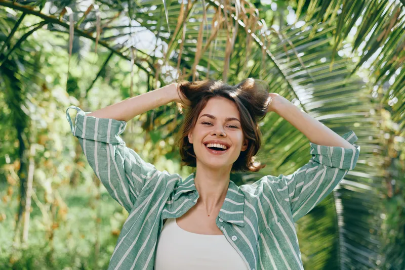 Joyful portrait of a smiling woman outdoors, under lush greenery, wearing a striped green shirt and white top, conveying carefree summer energy.