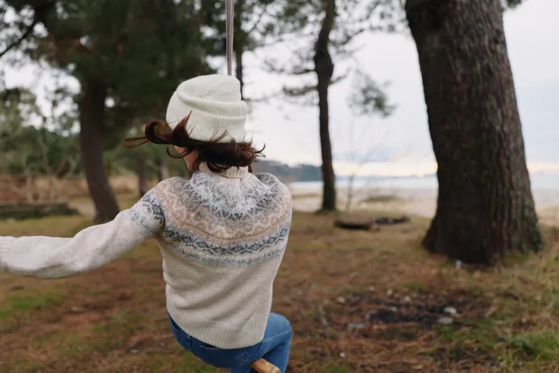 Outdoor scene of a woman spinning in a cozy sweater and beanie among trees, at a lake edge, capturing playful motion and fresh air in a serene park.