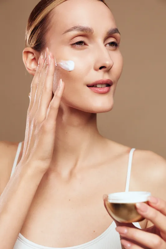 Young woman applying face cream on cheek, skincare routine, soft natural light, clear complexion, smooth skin texture, beige background, beauty concept, self care moment