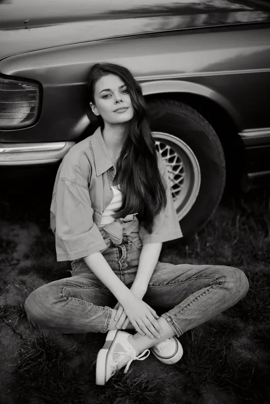 A young woman in casual attire poses confidently beside a vintage car in a serene black and white setting, conveying a sense of retro style and nostalgia.
