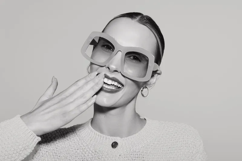 Smiling white woman with light skin wearing large sunglasses and knit sweater covers mouth with hand, studio portrait.